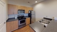 This single-floor unit offers a functional and well-organized layout, with distinct zones flowing naturally from one area to the next. The open-concept living area is finished with wall-to-wall carpet and features a ceiling fan, recessed lighting, and multiple windows that allow natural light to fill the space. A front entry door with adjacent windows further brightens the room, creating an inviting atmosphere.

The kitchen is efficiently designed with warm wood-toned cabinetry extending to the upper walls, providing ample storage. Dark granite-style countertops run along both sides of the kitchen, complementing the stainless steel appliances, which include a refrigerator, electric range, built-in microwave, and a double-basin sink. The kitchen floor features hardwood-style flooring, offering a clean contrast to the carpeted living areas.

The bathroom is well-appointed with a single-sink vanity topped with dark countertops, white cabinetry, a large mirror with overhead lighting, a full soaking tub with shower, and hardwood-style flooring. The bedroom is a generously sized, quiet space with neutral carpet and clean white walls, offering a blank canvas adaptable to various furnishing arrangements.