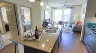 This single-story residence is designed around an open-concept layout that seamlessly connects the kitchen, dining, and living areas, creating a cohesive and functional living environment. The kitchen serves as a central focal point, anchored by a generously sized island topped with speckled granite countertops and fitted with a double stainless steel sink. Dark cabinetry beneath the island adds contrast, while pendant lighting overhead provides both task illumination and visual appeal.

Hardwood-style flooring extends throughout the main living areas, lending warmth and continuity to the space. The living area is filled with natural light from large windows dressed with horizontal blinds and sheer curtain panels, creating a bright and airy atmosphere. A ceiling fan is positioned overhead to ensure year-round comfort.

Adjacent to the main living space, a bedroom is visible through an open doorway, offering a glimpse of a well-lit, comfortably appointed sleeping area. Neutral wall tones throughout complement the overall design palette, making the space feel welcoming and versatile. The combination of modern finishes, thoughtful lighting, and open sightlines gives this residence a polished and livable character.