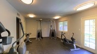 This single-floor fitness room is thoughtfully designed to accommodate a variety of workout routines. The space is finished with durable dark rubber flooring, which provides both cushioning and traction suitable for exercise. The room is equipped with a dual-cable resistance machine positioned centrally, making it a focal point for strength training. Cardio equipment, including an elliptical and a rowing or recumbent machine, is arranged along the perimeter, allowing for efficient use of the available floor space. Overhead, flush-mount ceiling lights provide consistent, even illumination throughout the room. A window on one wall allows natural light to enter, adding brightness and a connection to the outdoors. The walls are finished in a neutral tone, keeping the environment clean and distraction-free. The overall layout is practical and well-organized, making the space accessible and easy to navigate during workouts. This fitness room offers a self-contained environment suited for both cardio and strength training activities.