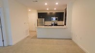This single-floor space features an open-concept layout that seamlessly connects the kitchen and living area, creating a bright and functional environment. The kitchen is outfitted with dark cabinetry that contrasts sharply against light-toned walls, lending a sleek, contemporary aesthetic. A granite-topped peninsula serves as a natural divider between the kitchen and living space, offering additional prep surface and casual seating potential. Stainless steel appliances, including a refrigerator, range, and over-the-range microwave, provide a polished, modern look. Recessed lighting throughout the kitchen ensures consistent, well-distributed illumination. The living area features neutral carpet flooring, contributing to a warm and comfortable atmosphere. Light-colored walls and recessed ceiling lights maintain a clean, airy feel across the open space. A full-panel entry door is visible from the main living area, positioned along a hallway that provides clear sightlines through the unit. The overall design balances practicality with modern style, offering a cohesive and move-in-ready environment suited for everyday living.