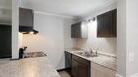 This single-story residence is designed with a practical, open layout that connects the kitchen and living area seamlessly. The kitchen is equipped with dark espresso cabinetry, light granite-style countertops, a double stainless steel sink with accent wall lighting, a coil-top electric range, a dishwasher, and a ventilation hood. A peninsula with a dark countertop separates the kitchen from the living space, providing a casual dining or workspace option. The living area features neutral carpet flooring, crown molding, a ceiling fan, and a door with sidelights that opens to a grassy outdoor area, allowing natural light to fill the space. A black refrigerator is positioned at the kitchen-living junction. The bedrooms are carpeted — one with darker carpet and a sliding-door closet with shelving, and another with lighter tones and similar closet storage. The bathroom area includes light hardwood-style flooring, a white vanity with a sink, bright mirror lighting, and a spacious sliding-door closet nearby. Textured ceilings and consistent crown molding carry throughout, lending a cohesive, well-maintained character to the entire unit.