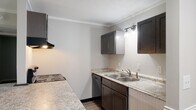 This single-story residence is designed with a practical, open layout that connects the kitchen and living area seamlessly. The kitchen is equipped with dark espresso cabinetry, light granite-style countertops, a double stainless steel sink with accent wall lighting, a coil-top electric range, a dishwasher, and a ventilation hood. A peninsula with a dark countertop separates the kitchen from the living space, providing a casual dining or workspace option. The living area features neutral carpet flooring, crown molding, a ceiling fan, and a door with sidelights that opens to a grassy outdoor area, allowing natural light to fill the space. A black refrigerator is positioned at the kitchen-living junction. The bedrooms are carpeted — one with darker carpet and a sliding-door closet with shelving, and another with lighter tones and similar closet storage. The bathroom area includes light hardwood-style flooring, a white vanity with a sink, bright mirror lighting, and a spacious sliding-door closet nearby. Textured ceilings and consistent crown molding carry throughout, lending a cohesive, well-maintained character to the entire unit.