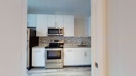 This single-floor space presents a cohesive, modern design throughout its layout. The kitchen is a standout feature, equipped with a full suite of stainless steel appliances, including a range, over-the-range microwave, and refrigerator. Sleek white flat-front cabinetry with bar-pull hardware lines the walls, complemented by light stone countertops and a distinctive mosaic tile backsplash in neutral tones that adds visual texture. A single-basin sink with a modern faucet completes the functional workspace.

Light hardwood-style flooring runs consistently through the main living areas and bedroom, contributing to the unit's bright, airy feel. The living area benefits from large windows fitted with horizontal blinds, allowing generous natural light to filter in while offering views of the surrounding neighborhood. Track lighting on the ceiling provides additional illumination. The bedroom includes a closet and connects to the main living space via a clean, open hallway.

The bathroom is finished with beige tile flooring and features a tub-shower combination enclosed by sliding glass doors. A wall-mounted vanity with dark wood cabinetry, a white countertop, and a framed mirror rounds out the space. Throughout, the clean white walls and consistent finishes create a unified, contemporary ambiance.