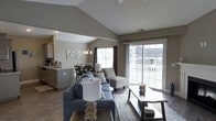 This single-story space showcases a thoughtfully designed open-concept layout that seamlessly connects the living, dining, and kitchen areas into one cohesive environment. The living area is anchored by a comfortable sectional sofa, accent chairs with patterned upholstery, and a low-profile wooden coffee table, all set atop a decorative area rug over rich hardwood flooring. A prominent vaulted ceiling rises above, lending an airy, spacious quality to the interior. Large sliding glass doors along the far wall flood the space with abundant natural light while providing direct access to an outdoor area, framing views of the surrounding neighborhood. The dining area features a round table with seating and is illuminated by a classic chandelier, creating an intimate yet open gathering space. Adjacent to the dining area, the kitchen is equipped with ample cabinetry, a sink, and counter workspace. Neutral taupe and gray tones throughout the walls and furnishings contribute to a calm, cohesive aesthetic, while recessed and pendant lighting provide warm illumination throughout the day and evening.