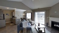 This single-story space showcases a thoughtfully designed open-concept layout that seamlessly connects the living, dining, and kitchen areas into one cohesive environment. The living area is anchored by a comfortable sectional sofa, accent chairs with patterned upholstery, and a low-profile wooden coffee table, all set atop a decorative area rug over rich hardwood flooring. A prominent vaulted ceiling rises above, lending an airy, spacious quality to the interior. Large sliding glass doors along the far wall flood the space with abundant natural light while providing direct access to an outdoor area, framing views of the surrounding neighborhood. The dining area features a round table with seating and is illuminated by a classic chandelier, creating an intimate yet open gathering space. Adjacent to the dining area, the kitchen is equipped with ample cabinetry, a sink, and counter workspace. Neutral taupe and gray tones throughout the walls and furnishings contribute to a calm, cohesive aesthetic, while recessed and pendant lighting provide warm illumination throughout the day and evening.