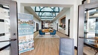 The Winslow Residences clubhouse at 13370 Magnolia Oak Circle in North Fort Myers, Florida, is a thoughtfully designed amenity facility spanning an expansive single floor. The grand entrance greets visitors with soaring ceilings highlighted by a dramatic arched skylight, designer pendant lighting, and wide-plank light wood flooring that flows throughout the space. The welcoming lobby transitions into an elegant lounge area furnished with a caramel leather sofa, teal accent chairs, and curated décor, anchored by a built-in beverage station along one wall. A second lounge features a bold circular ceiling feature, a large sectional, and floor-to-ceiling black-framed windows that flood the room with natural light and outdoor views. The games room is generously sized, accommodating a pool table, shuffleboard, and ping-pong table beneath a distinctive oval tray ceiling with gold accents, all bathed in natural light from oversized windows. A dedicated dining nook offers built-in banquette seating with marble-top tables and pendant lighting, ideal for casual gatherings. Private individual workspaces provide quiet, focused areas with built-in desks and garden views. A well-organized mail and package room features built-in mailboxes and patterned flooring. Additional nooks with textured feature walls and integrated shelving complete this vibrant, resort-inspired clubhouse.