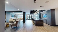 Located at 196 Westfield Rd in Noblesville, Indiana, this thoughtfully designed single-floor amenity space offers a generous, open layout that seamlessly blends social, recreational, and lounge areas. The kitchen area is a standout feature, anchored by a long, polished stone-topped island with seating for multiple guests outfitted in sleek black and gold chairs. Flanking the island are rich, dark wood cabinetry units with ample storage, a deep-basin sink, and a textured tile backsplash. A wall-mounted flat-screen TV is integrated directly into the kitchen backdrop, making it ideal for entertainment. The ceiling above showcases a dramatic coffered design with teal and dark accents, complemented by a mix of globe pendant lights and directional spotlights that add both warmth and visual interest. The living area transitions naturally from the kitchen, featuring light wood-look flooring, a textured accent wall, and large floor-to-ceiling windows that flood the space with natural light and provide views of an outdoor amenity area. A recreational zone includes a shuffleboard table beneath modern pendant lighting and additional wall-mounted screens. The lounge seating area offers comfortable arrangements of armchairs and sofas, creating an inviting, communal atmosphere throughout.