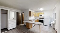 This single-floor space is designed with practicality and comfort in mind. The kitchen serves as the centerpiece, featuring light wood cabinetry that provides ample storage across both upper and lower cabinets. A built-in peninsula with a rounded countertop extension offers additional workspace and casual seating potential, making it a versatile hub for meal preparation and everyday use. The kitchen is fully equipped with a white appliance suite, including a refrigerator, electric range, built-in microwave, and dishwasher. A stainless sink is neatly integrated into the lower cabinetry, positioned beneath a layout that maximizes counter space. The flooring throughout transitions between wood-look planks, lending a warm, cohesive feel to the interior. Neutral white walls keep the space feeling bright and open, complemented by flush-mounted ceiling lighting that ensures even illumination. A solid wood door leads to an adjacent area, suggesting additional rooms or storage beyond the kitchen. The overall design prioritizes a clean, efficient layout that balances functionality with a welcoming, understated aesthetic suited to everyday living.