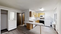 This single-floor space is designed with practicality and comfort in mind. The kitchen serves as the centerpiece, featuring light wood cabinetry that provides ample storage across both upper and lower cabinets. A built-in peninsula with a rounded countertop extension offers additional workspace and casual seating potential, making it a versatile hub for meal preparation and everyday use. The kitchen is fully equipped with a white appliance suite, including a refrigerator, electric range, built-in microwave, and dishwasher. A stainless sink is neatly integrated into the lower cabinetry, positioned beneath a layout that maximizes counter space. The flooring throughout transitions between wood-look planks, lending a warm, cohesive feel to the interior. Neutral white walls keep the space feeling bright and open, complemented by flush-mounted ceiling lighting that ensures even illumination. A solid wood door leads to an adjacent area, suggesting additional rooms or storage beyond the kitchen. The overall design prioritizes a clean, efficient layout that balances functionality with a welcoming, understated aesthetic suited to everyday living.