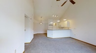 This single-floor space is defined by its dramatic vaulted ceilings that rise to an impressive peak, creating an expansive and airy atmosphere throughout the open-plan layout. The living area is finished with plush wall-to-wall carpet in a neutral gray tone, lending a comfortable and cohesive feel underfoot. Crisp white walls amplify the sense of space and brightness, reflecting light throughout the interior.

The kitchen is thoughtfully integrated into the open floor plan, separated by a wraparound breakfast bar that provides both a casual dining surface and a natural visual boundary. White cabinetry lines the kitchen walls, complemented by white appliances including a refrigerator and microwave, giving the space a clean and uniform aesthetic. A dedicated pendant light fixture illuminates the kitchen area, while a track lighting system extends across the vaulted ceiling to provide adjustable task and ambient lighting across the broader living space.

A ceiling fan with wooden blades is centrally mounted at the peak of the vault, ensuring efficient air circulation throughout the room. Additional pendant lighting contributes to the warm, welcoming ambiance. The overall design is clean, minimal, and highly functional, making the space feel open, versatile, and well-lit from every angle.