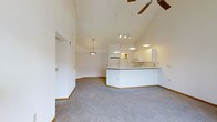 This single-floor space is defined by its dramatic vaulted ceilings that rise to an impressive peak, creating an expansive and airy atmosphere throughout the open-plan layout. The living area is finished with plush wall-to-wall carpet in a neutral gray tone, lending a comfortable and cohesive feel underfoot. Crisp white walls amplify the sense of space and brightness, reflecting light throughout the interior.

The kitchen is thoughtfully integrated into the open floor plan, separated by a wraparound breakfast bar that provides both a casual dining surface and a natural visual boundary. White cabinetry lines the kitchen walls, complemented by white appliances including a refrigerator and microwave, giving the space a clean and uniform aesthetic. A dedicated pendant light fixture illuminates the kitchen area, while a track lighting system extends across the vaulted ceiling to provide adjustable task and ambient lighting across the broader living space.

A ceiling fan with wooden blades is centrally mounted at the peak of the vault, ensuring efficient air circulation throughout the room. Additional pendant lighting contributes to the warm, welcoming ambiance. The overall design is clean, minimal, and highly functional, making the space feel open, versatile, and well-lit from every angle.