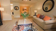 This single-floor residence offers a thoughtfully designed open-concept layout that seamlessly connects the living, dining, and kitchen areas into one cohesive and inviting space. The living room is anchored by a plush sofa accented with vibrant orange and jewel-toned throw pillows, centered around a marble-top coffee table resting on a bold, patterned area rug. A ceiling fan overhead enhances comfort throughout the space. Adjacent to the living area, the dining room draws the eye with a warm orange accent wall and an elegant chandelier that casts a soft, ambient glow over the dining table and chairs. The transition between rooms flows naturally, creating a sense of openness and continuity. A glimpse into the kitchen reveals cabinetry and appliances, completing the functional layout of the home. Neutral-toned carpeting throughout the main living areas adds warmth underfoot, while decorative mirrors and tasteful table lamps contribute to the space's polished, welcoming ambiance. The overall design balances comfort and style, making it a functional and visually appealing place to call home.