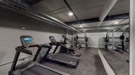 Located at 612 S Main St in Burlington, NC, this fitness facility offers a well-equipped, single-floor workout space designed for a comprehensive exercise experience. The space features an industrial aesthetic, with exposed silver HVAC ductwork running along the ceiling and recessed lighting that provides a focused, functional ambiance throughout.

The cardio zone is outfitted with multiple Star Trac treadmills and elliptical machines, each equipped with digital display consoles for tracking performance metrics. A large floor-to-ceiling mirror spans one wall, visually expanding the space and allowing users to monitor their form during workouts. Durable rubber flooring covers the entire area, providing traction and impact absorption suitable for a variety of training activities.

The strength training section includes a heavy-duty power rack complete with a barbell, weight plates, and an adjustable bench, accommodating a full range of resistance exercises. A dedicated free weights area features a well-stocked kettlebell rack with multiple weight options and a medicine ball rack, supporting functional and dynamic training routines. Additional equipment such as a rowing machine and step platforms further diversify the workout options available.

The space is organized to allow comfortable movement between zones, with clear sightlines across the floor plan. The overall design strikes a balance between utility and a modern, no-frills aesthetic suited to serious training.