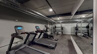 Located at 612 S Main St in Burlington, NC, this fitness facility offers a well-equipped, single-floor workout space designed for a comprehensive exercise experience. The space features an industrial aesthetic, with exposed silver HVAC ductwork running along the ceiling and recessed lighting that provides a focused, functional ambiance throughout.

The cardio zone is outfitted with multiple Star Trac treadmills and elliptical machines, each equipped with digital display consoles for tracking performance metrics. A large floor-to-ceiling mirror spans one wall, visually expanding the space and allowing users to monitor their form during workouts. Durable rubber flooring covers the entire area, providing traction and impact absorption suitable for a variety of training activities.

The strength training section includes a heavy-duty power rack complete with a barbell, weight plates, and an adjustable bench, accommodating a full range of resistance exercises. A dedicated free weights area features a well-stocked kettlebell rack with multiple weight options and a medicine ball rack, supporting functional and dynamic training routines. Additional equipment such as a rowing machine and step platforms further diversify the workout options available.

The space is organized to allow comfortable movement between zones, with clear sightlines across the floor plan. The overall design strikes a balance between utility and a modern, no-frills aesthetic suited to serious training.
