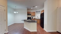 Located at 8439 Dorchester Rd in North Charleston, South Carolina, this single-floor residence offers a thoughtfully designed open-concept layout that seamlessly connects the kitchen and living areas. The kitchen is a functional centerpiece, featuring warm honey-toned cabinetry, dark granite countertops, and black appliances including a refrigerator, range, and microwave. A peninsula with a breakfast bar provides additional counter space and casual seating, making it ideal for both meal preparation and entertaining. Track lighting and a hanging pot rack add practical charm to the kitchen area. Rich hardwood-style flooring runs throughout the main living space, contributing to a warm and cohesive aesthetic. The adjacent dining area is highlighted by an elegant chandelier, creating an inviting atmosphere for formal or informal dining. Neutral wall tones throughout the space allow for versatile decorating options and help to amplify the natural light that flows through the unit. The open layout promotes an airy, connected feel across the living areas, making the space feel both comfortable and functional. The single-floor design ensures easy accessibility and a convenient flow between all areas of the home.