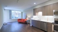 This single-floor space showcases a thoughtfully designed open-concept layout that seamlessly integrates the kitchen and living area. The kitchen is outfitted with contemporary flat-front cabinetry in a warm wood-tone finish, paired with a clean white countertop, a stainless steel sink with a modern pull-down faucet, and a stainless steel dishwasher. Recessed lighting illuminates the kitchen workspace, complemented by under-cabinet lighting that highlights the white tile backsplash. The living area benefits from a large picture window that invites abundant natural light and offers a pleasant view of mature trees and greenery outside. Rich, dark hardwood flooring runs throughout the space, providing a warm contrast to the light grey walls and white trim. Track lighting along the ceiling adds flexibility for illuminating the room. The overall ambiance is clean and modern, with a neutral palette that allows for versatile interior styling. The open layout and natural light create a sense of openness and airiness throughout the space.