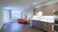 This single-floor space showcases a thoughtfully designed open-concept layout that seamlessly integrates the kitchen and living area. The kitchen is outfitted with contemporary flat-front cabinetry in a warm wood-tone finish, paired with a clean white countertop, a stainless steel sink with a modern pull-down faucet, and a stainless steel dishwasher. Recessed lighting illuminates the kitchen workspace, complemented by under-cabinet lighting that highlights the white tile backsplash. The living area benefits from a large picture window that invites abundant natural light and offers a pleasant view of mature trees and greenery outside. Rich, dark hardwood flooring runs throughout the space, providing a warm contrast to the light grey walls and white trim. Track lighting along the ceiling adds flexibility for illuminating the room. The overall ambiance is clean and modern, with a neutral palette that allows for versatile interior styling. The open layout and natural light create a sense of openness and airiness throughout the space.