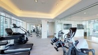 This sleek, contemporary fitness center is designed to offer a premium workout experience within a clean and modern environment. The space features a bright, open layout with a coffered ceiling accented by warm recessed lighting, creating an inviting and energizing atmosphere throughout.

The cardio zone is well-appointed with a variety of machines, including stationary bikes, ellipticals, stair climbers, and treadmills, all positioned to maximize usability and flow. Full-length floor-to-ceiling glass mirrors line one wall, enhancing the sense of space and allowing members to monitor their form during workouts. A mounted flat-screen display is integrated into the mirror wall, adding a modern, connected touch.

Beyond the main cardio area, the facility extends into a dedicated strength and multi-use training zone, equipped with cable machines and additional resistance equipment. Light hardwood-style flooring runs throughout, complementing the crisp white walls and contributing to the clean, minimalist aesthetic.

Natural light filters in through large windows, further brightening the space and creating an open, airy feel. The layout is thoughtfully organized, offering clear sightlines across the entire floor and ensuring a comfortable, distraction-free environment for all fitness levels.