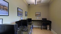 This single-floor computer room is designed as a functional shared workspace, offering multiple desktop computer workstations arranged along granite-patterned countertops that line the walls in an L-shaped configuration. Each station is equipped with a monitor, keyboard, and chair, providing individual users with dedicated workspace. The room features light-colored walls accented by three framed nautical-themed photographs, lending a calm and cohesive aesthetic to the space. Overhead, a drop ceiling with recessed fluorescent lighting ensures consistent, even illumination throughout the room. The light hardwood flooring complements the neutral wall tones, contributing to the space's clean and practical atmosphere. A security camera is mounted near the ceiling, indicating a monitored environment suited for semi-public or communal use. Informational printed notices are posted at select workstations, suggesting guidelines or instructions for users. The overall layout is efficient and straightforward, prioritizing accessibility and utility while maintaining a tidy, professional appearance appropriate for a business center, library, or residential amenity room.