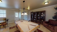 This dedicated billiards room offers a well-designed recreational space that combines classic game-room functionality with comfortable living elements. The centerpiece is a full-size pool table with an oak finish, positioned beneath an ornate wrought-iron pendant light fixture with dual frosted glass shades that provide focused illumination over the playing surface. The room features light tile flooring throughout, accented by decorative area rugs that define the game area and add warmth to the space.

Crown molding runs along the ceiling perimeter, lending a refined architectural detail to the room. A large, dark-wood built-in bookcase and entertainment unit lines one wall, offering ample storage and display space. A cozy armchair provides seating for spectators, while a wall-mounted television adds an entertainment dimension. Natural light enters through multiple windows fitted with horizontal blinds, and a set of French doors provides direct exterior access.

A separate dining nook within the space features a round glass-top table with ornate wrought-iron chairs, complemented by a framed wall art piece, making it a versatile area for casual dining or socializing. Multiple entry points, including a paneled door with a window, enhance accessibility and connectivity throughout the space.