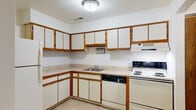 This two-floor residential space offers a practical and comfortable layout designed for everyday living. The kitchen is outfitted with an L-shaped configuration featuring white cabinetry trimmed in warm wood tones, ample upper and lower cabinet storage, a built-in dishwasher, an electric range, a refrigerator, and a double sink — providing a fully equipped cooking environment with easy-care tile flooring. A hallway connects key areas of the home, with a sliding closet providing additional storage near the entry to the kitchen. The spacious living areas are carpeted throughout, lending warmth underfoot, and feature large sliding glass doors that invite abundant natural light and offer views of the verdant, tree-lined surroundings outside. Additional carpeted rooms serve well as bedrooms, each with vertical-blind windows that frame leafy outdoor views. Two bathrooms are present: a full bath with a tiled tub-shower combination, mosaic tile flooring, and a vanity, as well as a half bath with a similar mosaic tile floor and vanity. A dedicated utility room houses the water heater, washer hookup, and electrical panel, ensuring all essential systems are conveniently accessible. The property is nestled within a green, tree-shaded setting visible from multiple vantage points throughout the home.