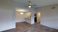 This single-story residence showcases an open-concept layout that seamlessly connects the living, dining, and kitchen areas. The living space features wood-look hardwood-style flooring that extends throughout, providing a warm and cohesive aesthetic. A ceiling fan with an integrated light fixture is centrally positioned, ensuring both airflow and ambient lighting across the main living area.

The dining area is defined by a stylish drum pendant light that hangs above, creating a designated gathering space adjacent to the kitchen. A pass-through counter with a built-in sink connects the dining area to the kitchen, offering both functional workspace and an interactive layout ideal for entertaining.

The kitchen is equipped with dark cabinetry, black appliances including a range, and overhead cabinetry providing ample storage. A separate entryway into the kitchen allows for ease of movement throughout the space.

The walls are finished in a neutral gray tone, complementing the flooring and contributing to a clean, modern ambiance. White trim and baseboards throughout add a crisp, polished contrast. The overall design prioritizes an efficient, open flow between functional areas.
