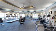 This fitness center is a comprehensively outfitted single-floor facility designed to support a full range of workout routines. The space is defined by its distinctive tray ceilings with teal accent banding, recessed lighting, and ceiling fans that contribute to an open, airy atmosphere. Large windows line multiple walls, flooding the interior with natural light and providing views of the surrounding exterior.

The cardio zone features a row of treadmills and elliptical machines from Precor, along with upright cycles and stair climbers, several equipped with individual display screens. A dedicated strength training area houses a multi-station cable machine and various selectorized weight machines, including a leg extension and press unit. Free weights are also available, with a full dumbbell rack offering a wide range of poundages neatly organized along one wall.

Floor-to-ceiling mirrors are strategically positioned throughout the space, visually expanding the room and allowing users to monitor their form. Wall-mounted televisions are installed at multiple viewing angles. The floor throughout is covered in durable blue-grey carpet. Additional accessories such as stability balls and medicine balls are stored along one wall, rounding out the facility's offerings.