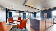 Located at 4066 Lexington Ave N in Shoreview, Minnesota, this sophisticated rooftop lounge offers a well-appointed living space designed with a modern, upscale aesthetic. The interior is defined by striking navy blue walls that provide a rich backdrop for warm hardwood flooring, creating an inviting yet refined atmosphere throughout.

The living room serves as the heart of the space, thoughtfully arranged with distinct seating areas. A cozy lounge nook features plush terracotta armchairs flanking a bold striped ottoman coffee table, with built-in wood shelving on either side adding both storage and character. Nearby, marble-topped bistro tables paired with tan leather chairs offer a more casual dining or gathering area adjacent to a sleek wet bar with dark, textured countertops.

A coffered ceiling detail with integrated linear lighting and a decorative metallic tile inset adds architectural interest and visual depth overhead. Large floor-to-ceiling windows line the perimeter, flooding the interior with natural light while framing expansive views of the surrounding Shoreview skyline and landscape.

The space connects seamlessly to a generously sized open-air terrace, finished with large-format stone tile and enclosed by modern glass-panel railings. String lights overhead and a built-in fire feature make the outdoor area ideal for year-round gatherings, offering unobstructed views of the neighborhood below.