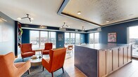 Located at 4066 Lexington Ave N in Shoreview, Minnesota, this sophisticated rooftop lounge offers a well-appointed living space designed with a modern, upscale aesthetic. The interior is defined by striking navy blue walls that provide a rich backdrop for warm hardwood flooring, creating an inviting yet refined atmosphere throughout.

The living room serves as the heart of the space, thoughtfully arranged with distinct seating areas. A cozy lounge nook features plush terracotta armchairs flanking a bold striped ottoman coffee table, with built-in wood shelving on either side adding both storage and character. Nearby, marble-topped bistro tables paired with tan leather chairs offer a more casual dining or gathering area adjacent to a sleek wet bar with dark, textured countertops.

A coffered ceiling detail with integrated linear lighting and a decorative metallic tile inset adds architectural interest and visual depth overhead. Large floor-to-ceiling windows line the perimeter, flooding the interior with natural light while framing expansive views of the surrounding Shoreview skyline and landscape.

The space connects seamlessly to a generously sized open-air terrace, finished with large-format stone tile and enclosed by modern glass-panel railings. String lights overhead and a built-in fire feature make the outdoor area ideal for year-round gatherings, offering unobstructed views of the neighborhood below.