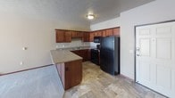 This single-floor residential unit offers a practical and well-organized layout designed for comfortable everyday living. The kitchen is a standout feature, outfitted with rich medium-toned wood cabinetry extending to the ceiling for generous storage, a granite-toned countertop, a double sink, and black appliances including a refrigerator, electric range, and microwave. A peninsula counter extends into the living area, creating a natural divide between the kitchen and the main living space while providing additional prep and seating space. The tile flooring in the kitchen carries a stone-like appearance, lending a durable and cohesive aesthetic throughout the functional areas of the home. The living area features neutral-toned carpet and smooth walls with standard baseboard trim, offering a clean and versatile canvas. The bathroom is well-appointed with a full tub and shower combination, a curved shower rod, a toilet, and a vanity, all set on the same stone-look tile flooring found in the kitchen. A dedicated laundry room provides added convenience with a stacked front-loading washer and dryer unit alongside a water heater, keeping all utilities neatly contained. Throughout the unit, warm wood door finishes and consistent neutral wall tones create a unified and tidy interior presentation.