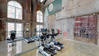 This fitness center occupies a striking adaptive reuse of a historic industrial building, blending raw architectural character with modern workout amenities. The space is defined by soaring ceilings with an open steel roof structure, exposed brick walls bearing original patina and vintage painted murals, and polished concrete floors that reflect the abundant natural light pouring in through oversized arched windows. Large glass doors at the far end of the main floor open to an outdoor area, further enhancing the bright, airy atmosphere.

The main workout floor is expansive, offering ample room for both cardio training and free-form exercise. A row of treadmills and elliptical machines by TRUE Fitness lines one wall, complemented by recumbent bikes for a well-rounded cardio selection. A dedicated free weights area features a full rack of dumbbells and a colorful assortment of kettlebells, suitable for strength and functional training. Yoga mats are available for stretching and floor work in the open central space.

A modern interior partition wall creates a corridor leading to additional rooms, finished in clean white and grey tones that contrast the industrial surroundings. Exposed conduit and ductwork run along the walls and ceiling, preserving the building's original industrial aesthetic throughout. The overall ambiance is energetic and distinctive, merging historic character with contemporary fitness functionality.