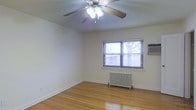 This single-floor space presents a straightforward and functional layout highlighted by several notable features. The room showcased in the image is finished with warm hardwood flooring that runs throughout, adding a timeless and classic character to the interior. The flooring is well-maintained and contributes a sense of warmth and continuity to the space.

A ceiling fan with an integrated light fixture is centrally mounted, providing both air circulation and ambient lighting. The walls are finished in a clean, neutral white tone, offering a versatile backdrop suitable for various décor styles.

Heating is provided by a traditional cast-iron radiator positioned beneath the window, a reliable and efficient system characteristic of well-established residential buildings. An additional wall-mounted air conditioning unit ensures year-round climate control. The window features horizontal blinds and allows natural light to filter into the room while offering a view of the surrounding outdoor environment.

The overall ambiance is clean, simple, and move-in ready, with practical amenities already in place. The space reflects a no-frills, functional design that prioritizes comfort and utility.