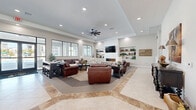 Located at 5353 N US Highway 27 in Ocala, Florida, this single-story clubhouse offers a generously sized, open-concept layout designed for both comfort and community engagement. The entry corridor features elegant travertine-style tile flooring with decorative inlay accents, flanked by glass-walled rooms that house a private office/business center and a seating area, all bathed in natural light from large windows.

The main lounge area is a standout, anchored by a shiplap-clad feature wall showcasing a wide linear electric fireplace, a wall-mounted flat-screen TV, and flanking built-in shelving units styled with greenery and décor. Rich leather seating, area rugs, and a tray ceiling with a modern ceiling fan contribute to the warm, inviting atmosphere.

A dedicated game room provides recreational options, complete with a full-size pool table, a granite-topped wet bar with bar seating, an additional wall-mounted TV, and a ceiling fan overhead. Bistro-style high-top tables line the wall of large black-framed windows, offering bright views of the surrounding community.

Throughout the space, large-format tile and carpet flooring define distinct zones, while recessed lighting and ceiling fans maintain a comfortable environment. The cohesive neutral palette and thoughtful furnishings create a welcoming, upscale communal setting.