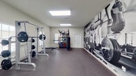 This fitness center offers a generously sized, single-floor workout space designed to support a comprehensive exercise regimen. The interior is bright and airy, benefiting from large windows fitted with white blinds that allow natural light to fill the room, complemented by flush-mounted overhead panel lighting throughout. The durable dark rubber flooring provides both safety and functionality across the entire space.

The cardio zone features multiple treadmills and elliptical machines, each positioned near windows to take advantage of natural light during workouts. A wall-mounted flat-screen television serves this area. The strength training section is equally well-appointed, housing a cable crossover machine with adjustable attachments, a multi-functional cable station, and a full power rack loaded with Olympic barbells and weight plates, accompanied by a flat bench. A dedicated free weights area includes a dumbbell rack and additional accessories.

A storage rack holds a variety of medicine balls, stability balls, and resistance bands, while a standing heavy bag rounds out the functional fitness offerings. One wall features a striking large-format black-and-white athletic mural that adds a motivational, gym-centric aesthetic to the space. A wall-mounted mirror further enhances the sense of openness. The overall ambiance is clean, modern, and purpose-built for serious training.