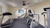 This dedicated fitness room is designed to support a well-rounded workout routine within a compact, efficiently organized space. The room features durable dark rubber flooring throughout, providing both cushioning and stability for exercise. Cream-colored walls and a textured ceiling contribute to a clean, neutral aesthetic, while track lighting mounted along the ceiling ensures consistent, bright illumination across the entire room. A ceiling fan adds air circulation, enhancing comfort during workouts.

The space is equipped with a notable selection of fitness equipment. Cardio options include two treadmills and an elliptical trainer, all from recognized brands including True Fitness. Strength training is supported by a Schwinn multi-station cable machine with a lat pulldown and adjustable bench, as well as a rack of dumbbells in graduated sizes. A large wall mirror spans one side of the room, visually expanding the space and allowing users to monitor their form.

A window on one wall allows natural light to filter in and provides a view of the surrounding exterior. A small storage rack near the entrance holds miscellaneous supplies. The room is accessed through a single door, keeping the space self-contained and purposeful.