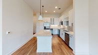 This well-appointed two-story home in Dallas, Texas offers a thoughtfully designed layout with modern finishes throughout. The kitchen serves as the heart of the home, showcasing a large central island with granite countertops, white shaker cabinetry with glass-front upper panels, stainless steel appliances, a built-in wine cooler, and warm hardwood flooring. Pendant lighting and recessed lights create an inviting atmosphere ideal for cooking and entertaining. The bedrooms feature soaring ceilings with ceiling fans and plush carpeting, lending a sense of volume and comfort. Natural light filters in through multiple windows fitted with blinds, offering views of the surrounding neighborhood. The bathrooms are finished with neutral tile flooring and walls, gray vanity cabinetry, and clean white countertops. One bathroom includes a frameless glass walk-in shower, while another offers a soaking tub. Each bedroom benefits from generous walk-in closet space with built-in shelving and hanging rods for ample storage. Additional rooms display rich hardwood flooring and ceiling fans, maintaining a consistent, contemporary aesthetic throughout both floors of the home.