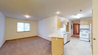 This single-floor space is designed with an open-concept layout that seamlessly connects the kitchen and living area. The kitchen is equipped with light oak cabinetry, both upper and lower, providing ample storage. A peninsula with a light-colored countertop extends outward, creating a natural divide between the kitchen and the carpeted living area while offering additional workspace or casual seating. A stainless steel sink is positioned along the back wall, accompanied by a white dishwasher and under-counter cabinetry. The kitchen features tile flooring, which transitions to wall-to-wall carpet in the living area. Recessed lighting and flush-mount ceiling fixtures provide consistent illumination throughout the space. A window in the living area allows for natural light to enter. The textured ceiling adds a subtle design element overhead. A hallway visible at the rear of the space leads to additional rooms, suggesting a functional layout beyond the main open area. The overall aesthetic is clean, practical, and neutral, with warm wood tones and light walls contributing to a bright, welcoming atmosphere.