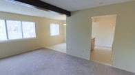 This single-floor space offers an open, airy layout with a seamless flow between the main living area and an adjoining dining or secondary room. The interior is finished with soft, wall-to-wall carpeting throughout, creating a warm and comfortable atmosphere underfoot. Neutral, light-toned walls reflect natural light generously, contributing to the overall brightness of the space.

The main living area is illuminated by a wide bank of windows dressed with vertical blinds, allowing for flexible light control while maintaining a clean, classic look. A textured ceiling adds a subtle visual element to the expansive overhead space. A wrought iron railing detail is visible, hinting at a split-level or elevated entry design that adds architectural character.

The adjoining room, accessible through a wide open archway, features its own window with vertical blinds and is accented by an elegant chandelier overhead, lending a touch of sophistication. The open-plan design between these two spaces promotes a sense of generous square footage, making the interior feel connected and easy to navigate.

Overall, the space presents a clean, neutral palette that is move-in ready and well-suited for a variety of furnishing styles, with natural light and an open layout as its defining features.