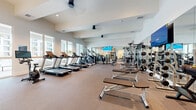 Located at 1799 North Williamson Boulevard in Daytona Beach, FL, this single-floor fitness center is thoughtfully designed to support a wide range of workout routines. The facility is divided into two distinct exercise rooms, together offering a comprehensive and well-organized training environment.

The main exercise room is spacious and bright, featuring large windows along one wall that flood the space with natural light and provide outdoor views of the surrounding landscape. The room is lined with full-length mirrors that enhance the sense of openness. Cardio equipment — including multiple treadmills, ellipticals, and a stationary bike — is arranged along the windowed wall, while the strength training section houses a robust selection of machines, including Life Fitness and Hammer Strength cable systems, weight racks, and adjustable benches. Wall-mounted flat-screen TVs add an entertainment element to workouts. The elevated ceiling with a coffered detail adds architectural character to the room.

The secondary exercise room offers a more intimate setting with light-toned carpet, recessed lighting, and clean white walls. It features a dedicated cycling station with a screen-equipped bike, a full dumbbell rack with mirror backing, and a boxing area complete with a heavy bag and wall-mounted training rack — ideal for high-intensity interval and combat-style training.
