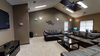 This single-story space centers around a spacious living room defined by striking vaulted ceilings that lend an open, airy feel to the interior. The dramatic pitch of the ceiling is accentuated by multiple skylights, which draw in an abundance of natural light and brighten the room throughout the day. A ceiling fan is mounted at the apex, providing both functionality and a visual anchor to the soaring roofline.

The living area is finished with warm, taupe-toned walls that create a cozy and inviting atmosphere, complemented by plush, neutral-toned carpeting underfoot. The seating arrangement features dark leather sofas paired with a rich, dark-stained wood coffee table, lending the space a grounded, contemporary aesthetic. Decorative wall art adds a subtle artistic touch to the room's overall character.

A small desk area with a computer monitor is tucked toward the back corner, offering a functional workspace within the living space. Additional wall-mounted light fixtures supplement the natural light from above, ensuring the room remains well-lit at any hour. The layout feels generous and well-proportioned, balancing comfort with visual openness.