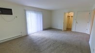This single-floor space presents a clean, functional layout centered around a generously sized carpeted living area. The room is finished with neutral-toned carpet and light-colored walls, creating a bright and open atmosphere. A large sliding glass door or wide window dressed with vertical blinds dominates one wall, allowing substantial natural light to fill the space while offering potential outdoor access or views. A baseboard heating unit runs beneath the window, providing an efficient heating solution. The open doorway visible on the opposite side leads to a walk-in closet, complete with a closet rod and shelving, offering practical storage capacity. The overall design is straightforward and understated, with smooth white ceilings and minimal architectural detailing that lend the space a versatile, move-in-ready quality. The neutral finishes throughout make it easy to personalize with furnishings and décor. The layout appears efficient, with the living area connecting conveniently to the storage space, supporting comfortable day-to-day functionality.