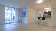 This single-floor space offers a clean, contemporary aesthetic characterized by an open-plan layout that promotes a sense of spaciousness and easy flow between areas. Light-toned tile flooring runs throughout, reflecting natural light and contributing to the space's airy, bright atmosphere.

The living and dining area is anchored by large floor-to-ceiling windows and a sliding glass door that floods the room with abundant natural light and provides views of lush tropical greenery outside. A sleek, round white dining table paired with modern acrylic chairs adds a minimalist touch, while a vibrant blue ocean-themed artwork on the wall serves as a striking focal point.

Adjacent to the main living area, a bar-height counter with contemporary black bar stools defines the dining or breakfast space, illuminated by a stylish pendant light that adds warmth and visual interest. Smooth, neutral-toned walls and white baseboards maintain a cohesive, understated palette throughout.

The layout transitions naturally through a gated doorway into further interior areas, hinting at additional rooms beyond. Recessed and flush-mount ceiling lighting ensures the space remains well-lit at all times. Overall, this residence presents a light-filled, modern environment with a relaxed yet polished character, well-suited to both everyday living and entertaining.