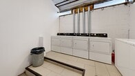 Located at 500 Avenue des Pins Ouest in Montréal, this shared laundry facility is a compact, functional space designed to serve building residents efficiently. The room is finished with white-painted concrete block walls and light-toned ceramic tile flooring, giving it a clean, utilitarian appearance. A slight raised platform along the perimeter of the room positions the machines for convenient use.

The facility is well-equipped, featuring multiple front-loading washing machines and dryers arranged along two walls, providing ample capacity for shared use. The machines are modern units with digital control panels, and the dryers are fitted with stainless steel exhaust vents that vent through the exterior wall. A centrally mounted electronic payment terminal allows residents to operate the machines via a card or payment system, eliminating the need for coins.

A small window near the ceiling provides a modest amount of natural light, while exposed utility piping and ductwork along the ceiling reflect the practical, no-frills nature of the space. A single white door with posted building notices serves as the main point of entry. A floor drain is integrated into the tiled floor, ensuring proper water management. Overall, the space is straightforward and well-organized, prioritizing functionality and ease of use for its residents.