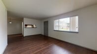 This single-floor, two-bedroom, one-bathroom unit offers a clean and functional layout designed for comfortable everyday living. The open living area serves as the heart of the home, featuring rich, dark hardwood-style flooring that extends throughout the main spaces. A large window with horizontal blinds fills the room with natural light and provides views of the surrounding exterior. A pass-through opening connects the living area directly to the kitchen, adding an open and communicative feel between the two spaces.

The galley-style kitchen is efficiently organized, with dark cabinetry lining both sides of the corridor, complemented by light wood-style flooring. Black appliances, including a refrigerator, dishwasher, and electric range, are neatly integrated alongside a single-basin sink, providing all essential cooking and meal preparation amenities.

Both bedrooms are carpeted in a neutral tone, offering a softer, quieter environment. Each room includes a window that admits natural light and provides outdoor views, along with ceiling-height ventilation and ample wall space for furnishings.

The bathroom features a full tub-shower combination dressed in large-format white subway tile with a contrasting accent band, chrome fixtures, and a small window for natural ventilation—creating a bright, clean aesthetic throughout.