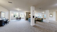 Located at 1745 Graham Ave in Saint Paul, MN, this generously sized single-floor community lounge offers a welcoming and versatile gathering space. The main lounge area is anchored by elegant white structural columns and features comfortable seating arrangements centered around a fireplace, creating a warm and inviting atmosphere. Patterned carpeting flows throughout, complementing the neutral, soft color palette of the walls and furnishings. Large windows along the perimeter and soaring two-story glazing in a sunlit corner nook flood the space with natural light and offer pleasant views of surrounding greenery, enhancing the open, airy feel. A dedicated card and game area accommodates multiple seating groups with classic wood and upholstered chairs. Private dining rooms provide intimate settings for smaller gatherings, each outfitted with formal dining tables and garden-facing windows. The fully equipped kitchen features warm wood cabinetry, white countertops, a stone tile backsplash, and stainless steel appliances including a refrigerator, range, microwave, and dishwasher, making the space well-suited for catered events or community meals. The overall design balances functionality with a comfortable, residential ambiance throughout.