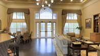 This single-story space showcases a grand, open living area defined by its impressive architectural details and refined aesthetic. The room's most striking feature is its soaring ceiling height, which creates an expansive, airy atmosphere enhanced by an ornate chandelier that serves as a central focal point. Tall, arched transom windows flank a set of elegant French doors, flooding the space with natural light while offering views of the lush greenery outside. The windows are dressed with decorative valances and floor-length drapes in warm gold tones, adding a layer of sophistication to the overall design. Dark-stained hardwood flooring anchors the room, providing a rich contrast to the soft, neutral wall tones. The living area is thoughtfully arranged with a plush sofa, upholstered accent chairs, and a traditional area rug, creating an inviting and comfortable seating arrangement. A dining area is also visible to the left, offering a functional yet stylish complement to the living space. Crown molding and classic architectural trim further enhance the formal, traditional character of this well-appointed interior.