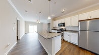 This well-appointed single-story home showcases a thoughtfully designed open-concept layout with hardwood-style flooring flowing seamlessly throughout. The kitchen serves as the heart of the home, featuring a large central island with granite countertops and an integrated sink, crisp white cabinetry, a classic subway tile backsplash, and a full suite of stainless steel appliances including a gas range, over-the-range microwave, and refrigerator. Pendant lighting above the island adds a refined touch, while crown molding along the ceilings lends an elegant finish to the space. The kitchen opens directly into the living area, which is bathed in natural light from a sliding glass door that leads to an outdoor patio. The bedrooms offer a calm, neutral palette with generous closet space, including a walk-in closet with built-in shelving and hanging rods. The bathrooms are clean and functional, each featuring a full soaking tub with a tiled surround and modern chrome fixtures. Additional highlights include recessed lighting, ceiling fans, and a dedicated pantry with open shelving. The overall ambiance is bright, clean, and move-in ready, presenting a comfortable and practical living environment.