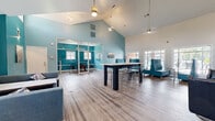 This single-floor workspace is thoughtfully designed to accommodate a variety of working styles, combining open collaborative areas with private enclosed offices. The main open area is defined by striking vaulted ceilings that dramatically increase the sense of volume, complemented by pendant lighting and ceiling fans with a gold finish. Light wood-tone flooring runs consistently throughout the space, unifying the layout visually.

The dominant design element is a bold teal accent color applied to walls and interior partitions, creating a vibrant yet professional atmosphere against the neutral white ceiling and trim. The open coworking area features a tall communal bar-height table with metal stools, ideal for group work or casual meetings. Additional seating options include modern teal booth-style sofas and lounge chairs positioned near windows that allow natural light to filter in.

Glass-walled, aluminum-framed private offices are accessible from the main area, offering visual transparency while maintaining acoustic separation. Inside, these offices include individual desks and seating. A dedicated computer workstation area with multiple desktop setups and acrylic chairs is also present. A compact entry or transition area features a curved modern bench seat. The overall ambiance is bright, contemporary, and well-suited for productivity.