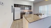 This single-story residence offers a thoughtfully designed open-concept layout that seamlessly connects the living and kitchen areas. The generous living room is finished with neutral wall-to-wall carpeting and benefits from abundant natural light through large windows dressed with vertical blinds. A ceiling fan adds both comfort and function to the space.

The kitchen is well-equipped, featuring dark wood-toned cabinetry contrasted by granite-style countertops and a peninsula with a built-in sink, providing additional workspace and a natural division between the kitchen and living area. White appliances, including a refrigerator, electric range, microwave, and dishwasher, are all included. Light wood-look flooring runs throughout the kitchen, creating a clean, practical surface.

The bedrooms are carpeted and filled with natural light from multiple windows with vertical blinds. One bedroom includes direct access to a bathroom, which features a granite-topped vanity, combination tub and shower, and light wood-look flooring. A walk-in closet with wire shelving provides ample storage. A second bathroom with a similar granite vanity serves the remaining rooms. The overall ambiance is clean, bright, and move-in ready.