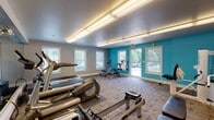 The fitness center at 3 Schooner On in Milford, Connecticut, is a well-equipped, single-floor facility designed to support a comprehensive workout experience. The space is defined by its bold teal accent walls contrasted against neutral gray tones, creating an energetic and motivating atmosphere. Flush-mounted fluorescent lighting runs across the ceiling, providing consistent illumination throughout the room, while multiple windows along the perimeter allow ample natural light and outward views of the surrounding greenery.

The layout is thoughtfully organized to accommodate a variety of fitness activities. The cardio zone features multiple treadmills, elliptical trainers, stationary bikes, and a rowing machine, all arranged to maximize space and usability. A multi-station cable weight machine anchors the strength training area, complemented by a full rack of dumbbells in varying weights, kettlebells, and additional free-weight accessories. A wall-mounted mirror expands the sense of space and supports form-checking during workouts.

The free-movement area includes a exercise mat and a BOSU balance trainer, ideal for stretching, core work, and functional training. A wall-mounted television adds entertainment options for users. Carpeted flooring with a patterned tile design covers the entire space, offering comfort underfoot. A direct exterior door provides convenient access to the outdoors.